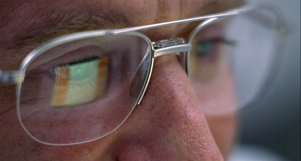One Hour Photo (2002) movie still: closeup — Close-up of a person's eyes behind wire-rimmed glasses in a dim environment. A computer screen's…