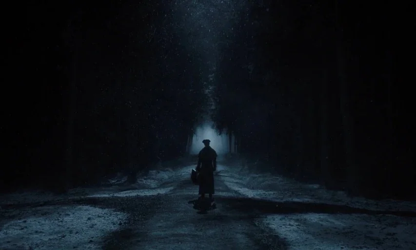 Nosferatu (2024) movie still: wide — A lone figure stands on a snow-covered dirt path flanked by dark trees. Nighttime scene with blue moonlight…