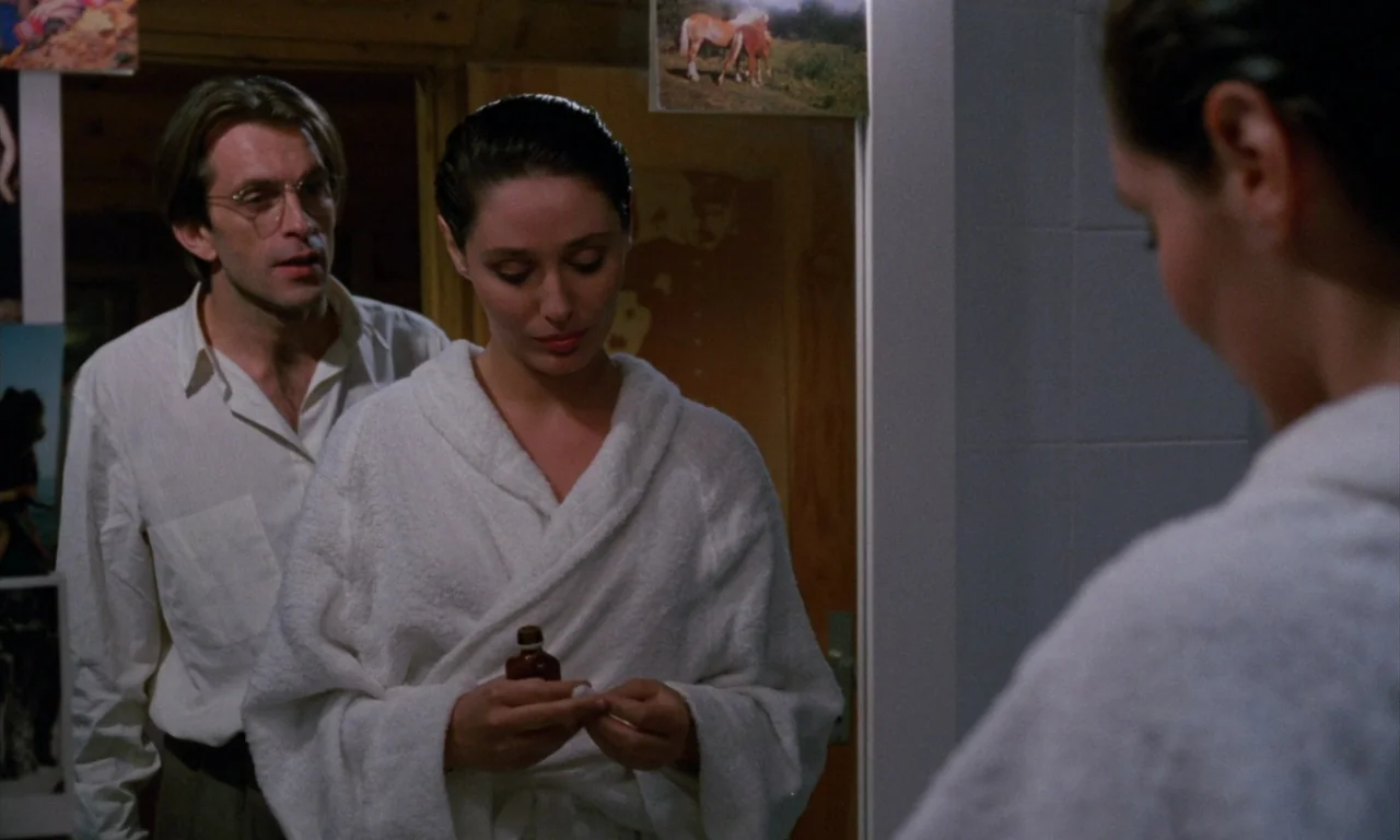 The Church (1989) movie still: medium — A woman in a white bathrobe stands facing a mirror in a dimly lit area. Beside her, a man in a white dress…