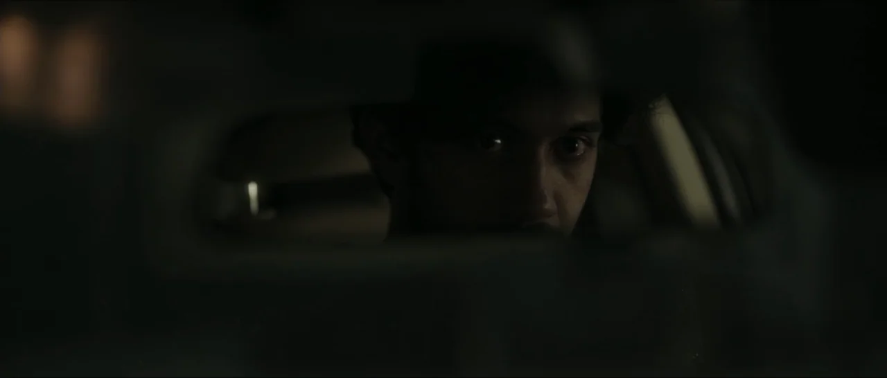 Innocent Vengeance (2023) movie still: closeup — A dark car interior seen through the reflective glass of a rearview mirror. A man stares into the…