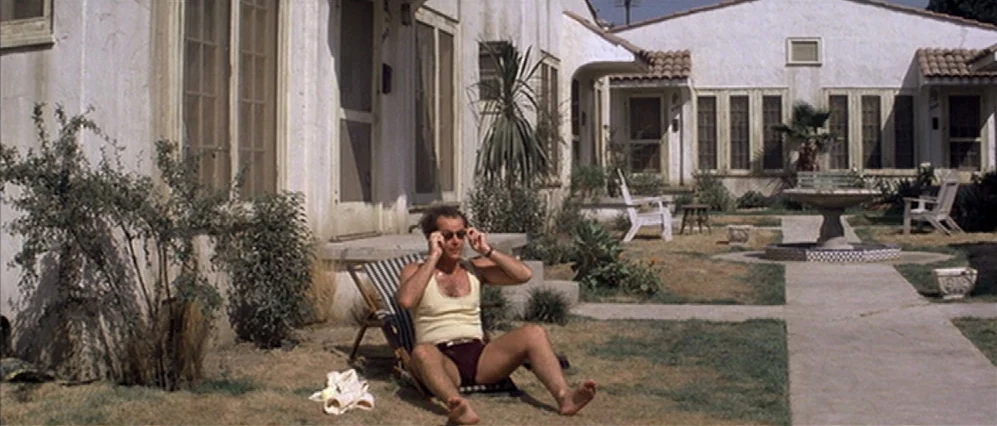 The Fortune (1975) movie still: fullbody — A sunny residential courtyard featuring white stucco buildings with tiled roofs and dry grass. A man sits…
