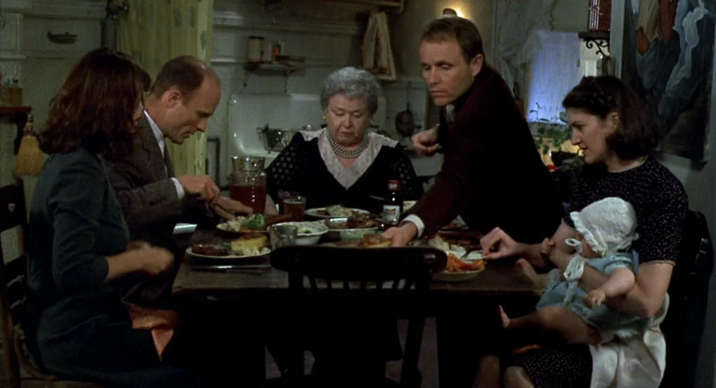 Pollock (2000) movie still: medium — A group of five adults and an infant sit at a wooden dinner table. They are eating a meal together inside a…