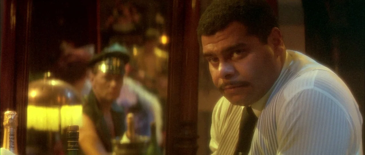 Querelle (1982) movie still: medium — A dim bar interior with dark wood paneling and gold-toned light accents. A man in a striped dress shirt looks…