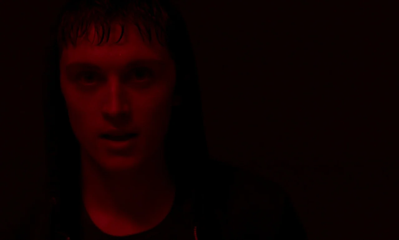 It Is In Us All (2022) movie still: closeup — A dark, featureless environment illuminated by a single red light source. A young man faces forward…