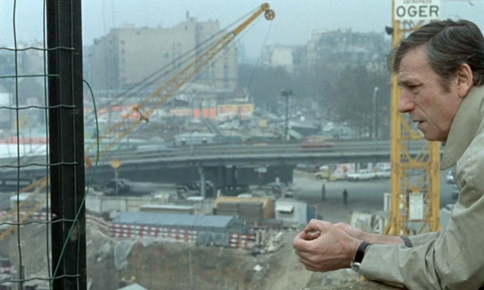 Tout Va Bien (1972) movie still: medium — An urban construction site with a yellow crane sits beneath a grey, overcast sky. A middle-aged man in a…