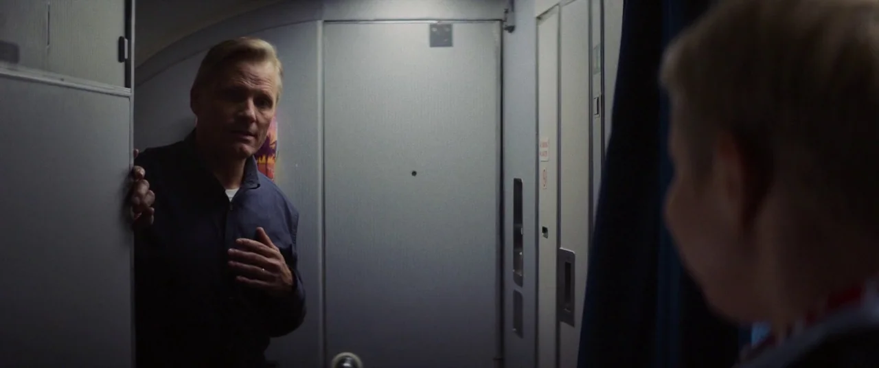 Falling (2020) movie still: medium — The narrow aisle of a commercial aircraft shows a gray lavatory door. A man in a dark shirt stands by the door…