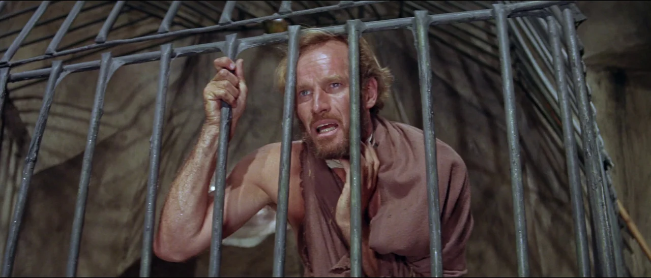 Planet of the Apes (1968) (1968) movie still: medium — A man sits confined within a set of heavy, dark metal bars. He grips the cold metal structure…