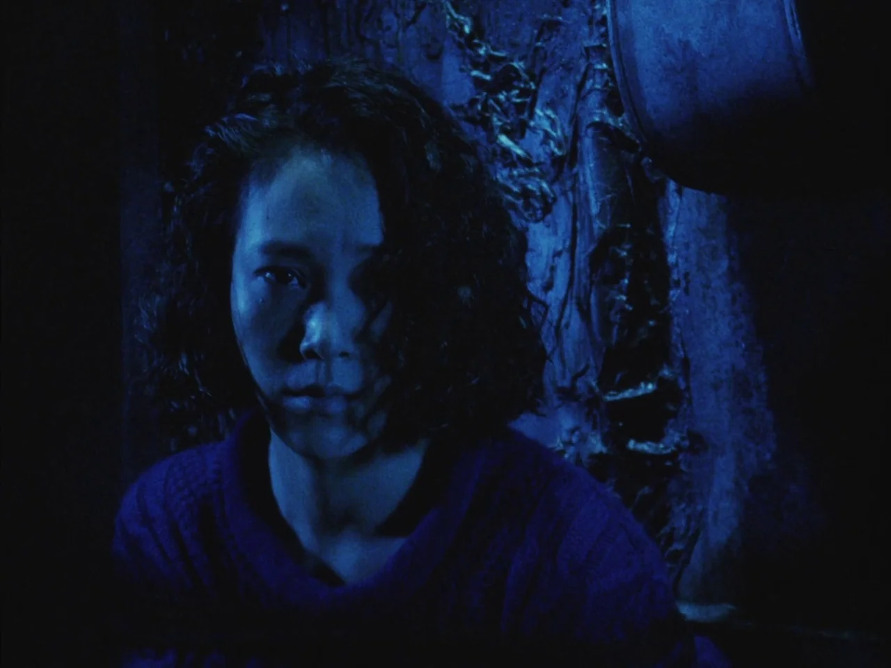 Tetsuo II: Body Hammer (1992) movie still: closeup — A person stands against a dark, textured wall with organic-looking relief details. She faces…