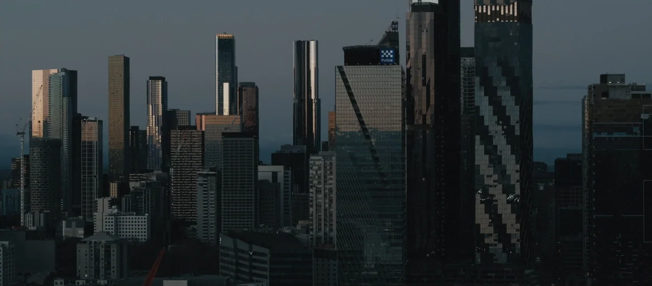 Force of Nature: The Dry 2 (2024) movie still: establishing — A dense urban cityscape features numerous glass skyscrapers rising against a darkening…