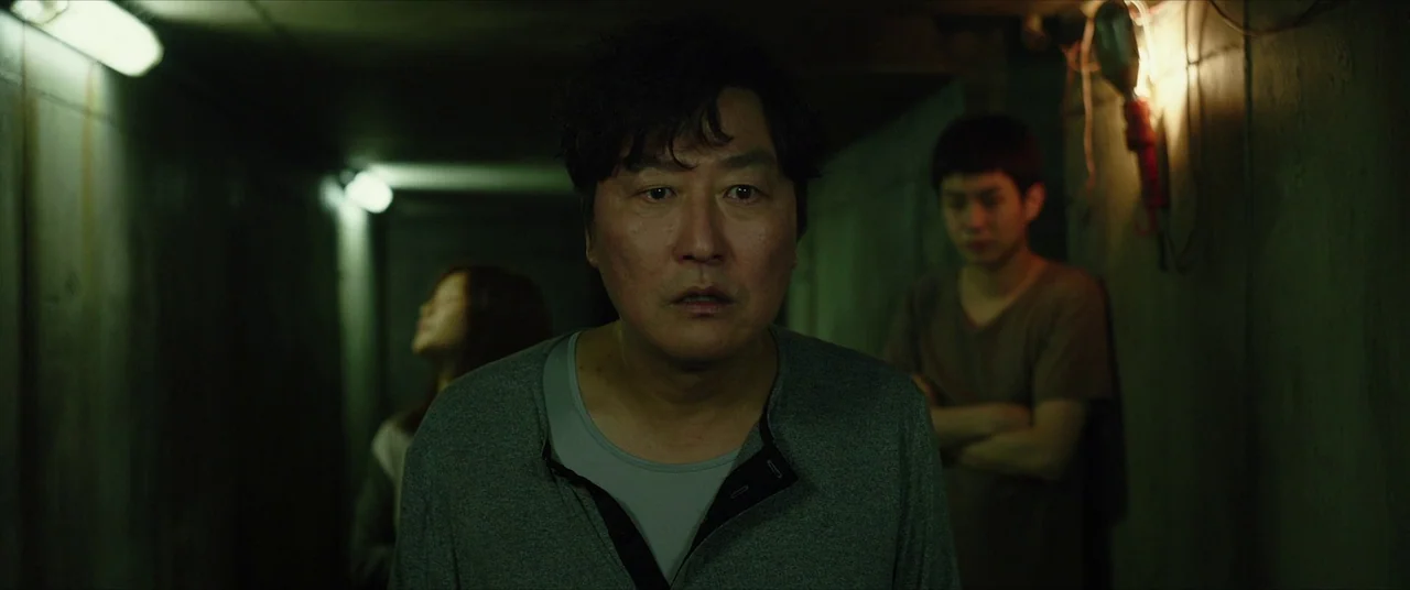 Parasite (2019) movie still: medium — A dimly lit, narrow concrete corridor with visible wiring and industrial wall textures. A middle-aged man…