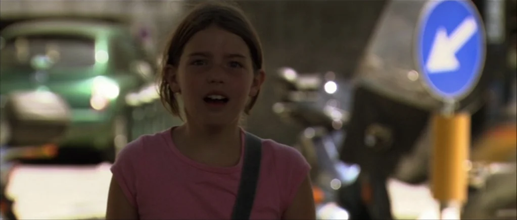 Genova (2008) movie still: medium — A girl stands on a city street with a green car and traffic sign nearby. She is looking forward with her mouth…