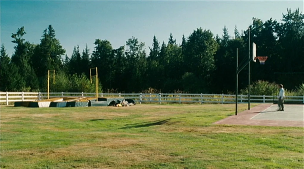 Visioneers (2008) movie still: wide — A flat field features a high jump mat and a basketball hoop structure. A lone man stands on the paved…