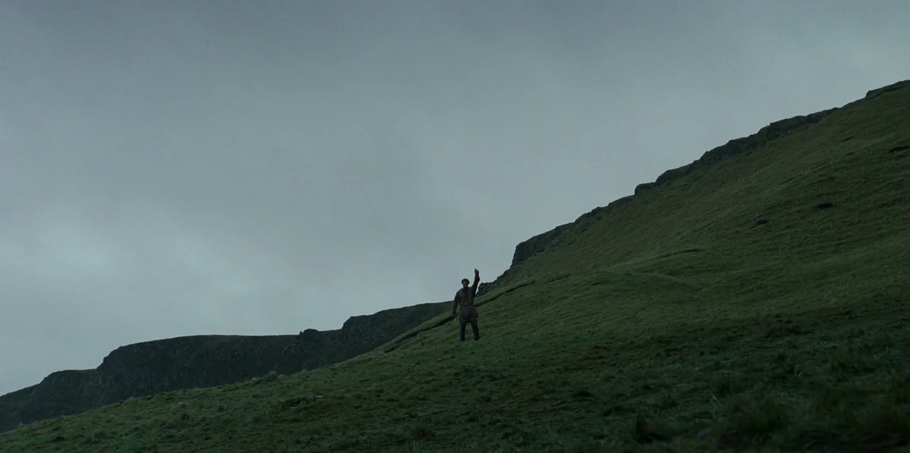The Northman (2022) movie still: wide — A vast, grassy hillside under a heavy, grey, overcast sky. A lone man stands on the incline holding one arm…