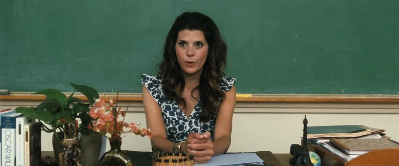Crazy, Stupid, Love. (2011) movie still: medium — A teacher sits at a wooden desk with books and plants before a chalkboard. A woman in a blue…