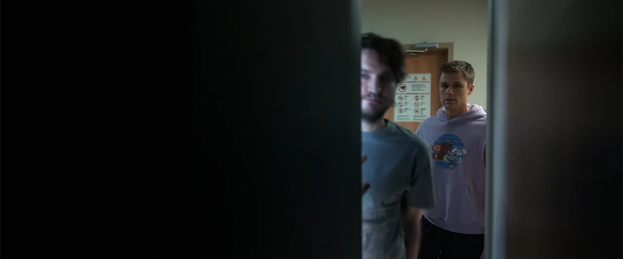 Final Destination Bloodlines (2025) movie still: medium — A hospital hallway is partially visible through an open door frame. Two young men stand in…