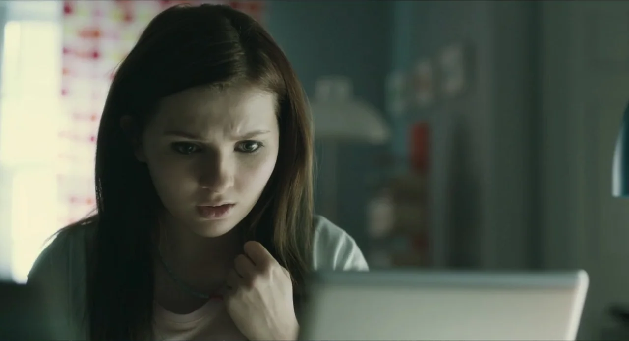 Haunter (2013) movie still: closeup — A dimly lit bedroom with a window in the background featuring patterned curtains. A teenage girl looks…