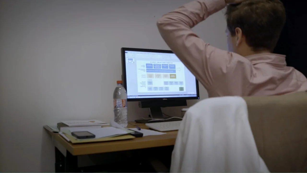 Brexit: Behind Closed Doors (2019) movie still: medium — A cluttered desk workspace sits before a plain wall inside a quiet room. A person sits at…