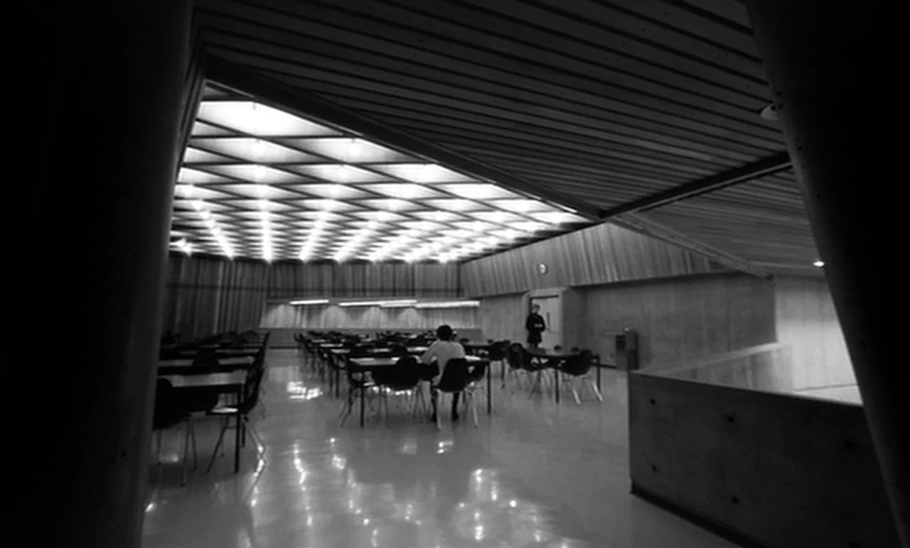 Stereo (1969) movie still: wide — A large cafeteria space features a geometric grid ceiling and polished reflective floors. A lone individual sits…
