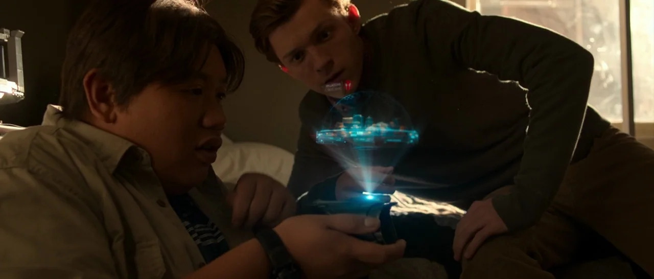Spider Man: Homecoming (2017) movie still: medium — A dimly lit room contains two teenagers observing a glowing blue holographic interface. They…