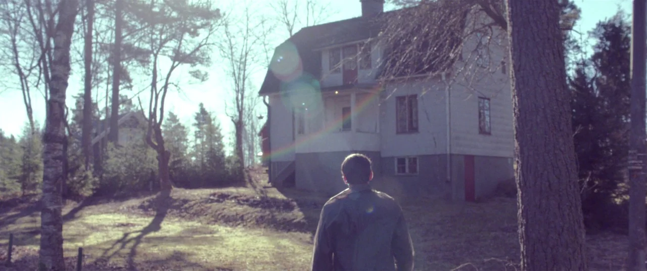 The House That Jack Built (2018) movie still: medium — A man walks across a grass lot toward a two-story wooden house. Tall trees surround the house…