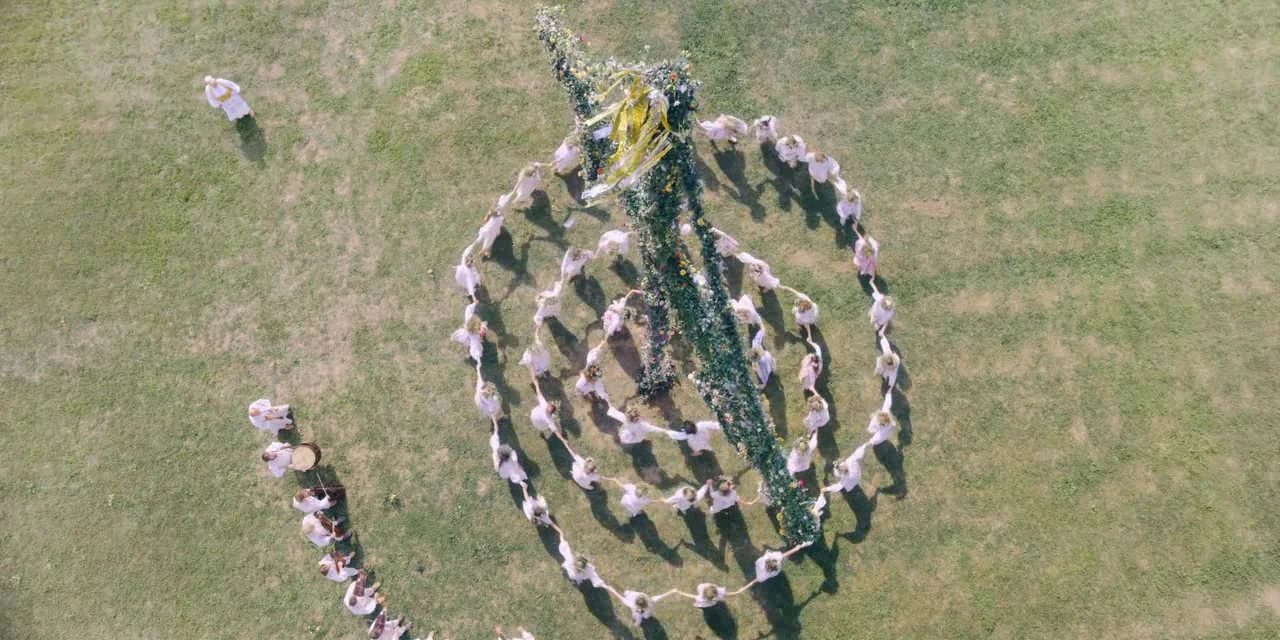 Midsommar (2019) movie still: wide — A vast, open grassy field contains a tall, flower-covered maypole at the center. Dozens of people wearing white…