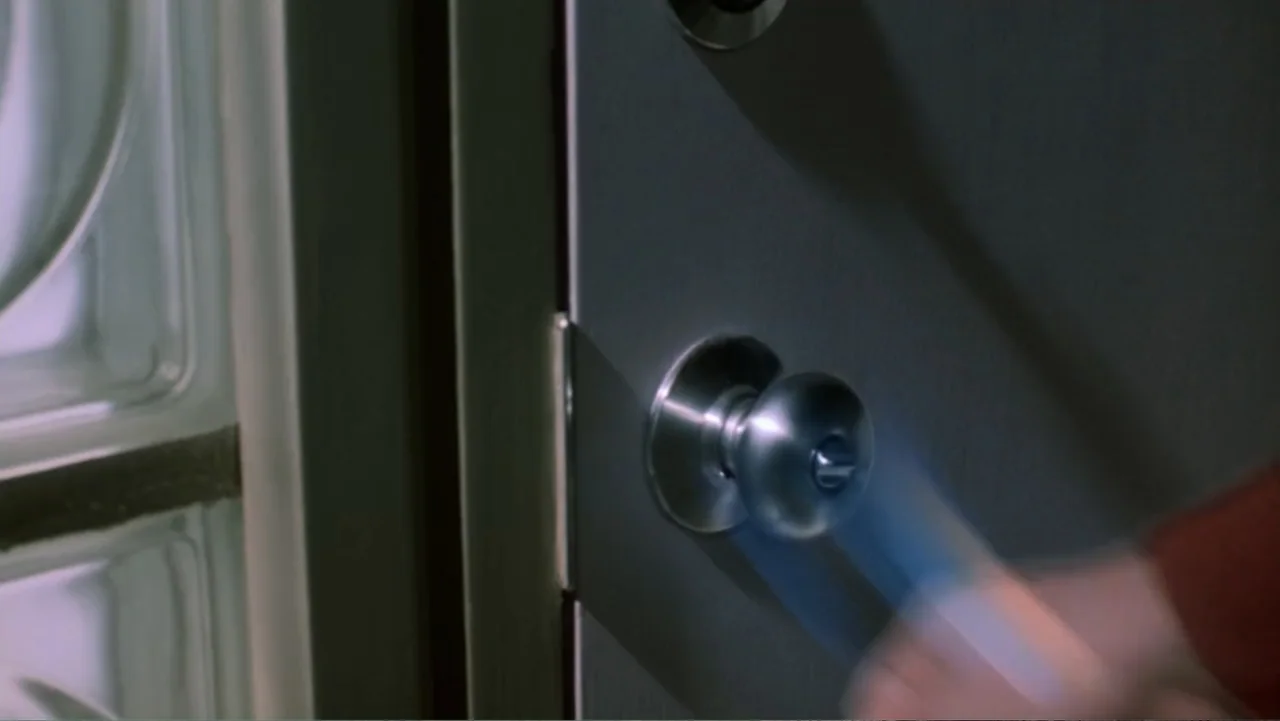 Ghost (1990) movie still: closeup — A close-up shot focuses on a metal door knob attached to a grey door. A blurred hand moves to grip the door knob…