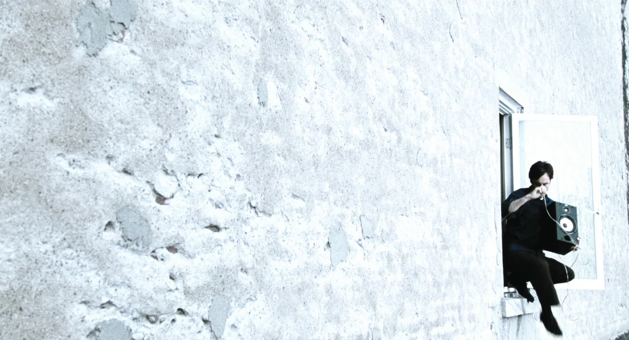 Last Night (1998) movie still: wide — A man sits on a stone window ledge against a rough, weathered plaster wall. He holds a single black speaker…