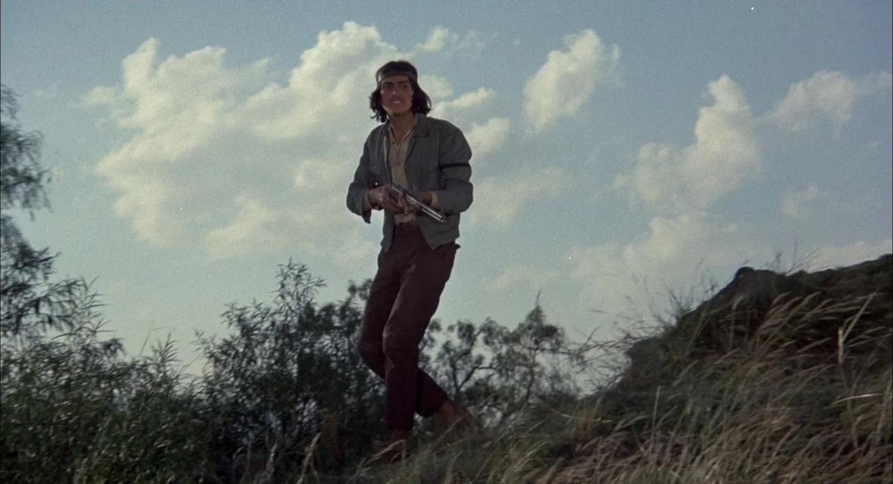 Bring Me The Head of Alfredo Garcia (1974) movie still: fullbody — A man stands on a sloping grassy hill against a bright sky with light clouds. He…