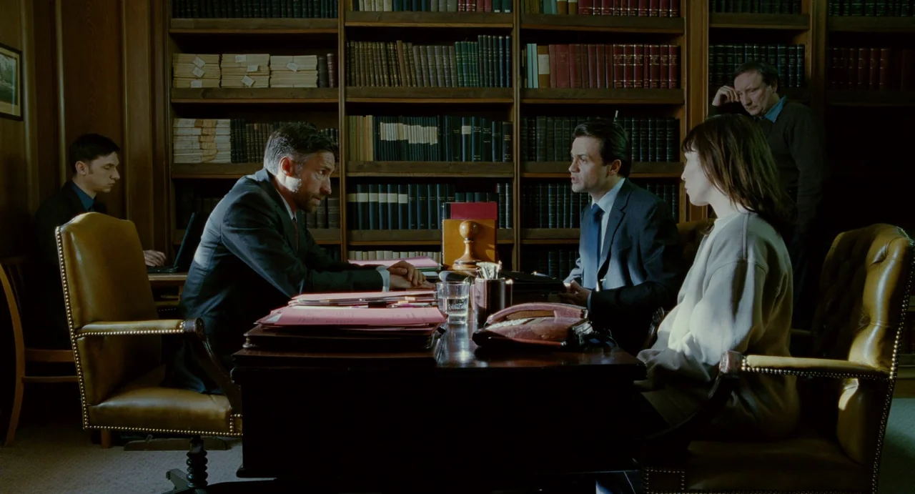 Passion (2012) movie still: medium — A wood-paneled room lined with full bookshelves and vintage leather chairs. A group of professionals sit around…