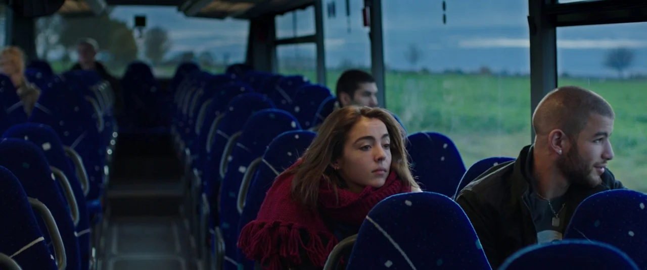 Raw (2016) movie still: medium — A quiet commuter bus interior with rows of blue fabric seats in dim light. Two young passengers sit near each other…