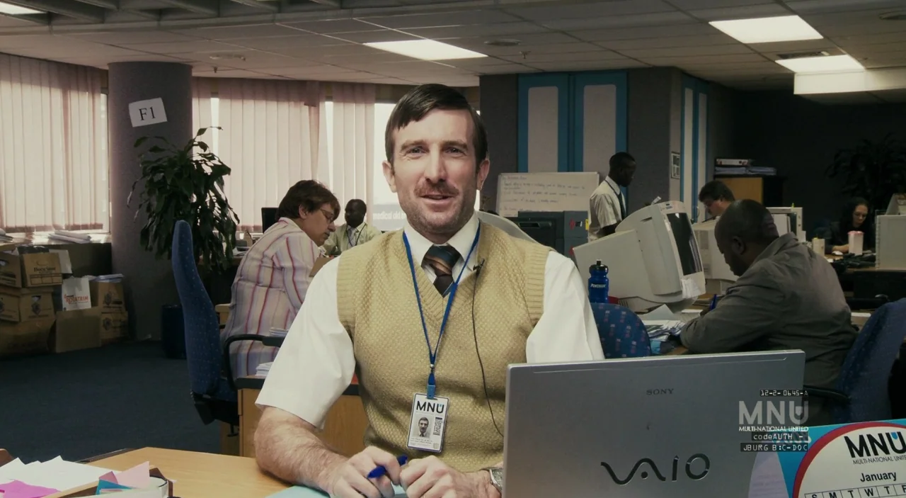 District 9 (2009) movie still: medium — A busy open-plan corporate workspace features cubicles and multiple employees working at desks. A man…
