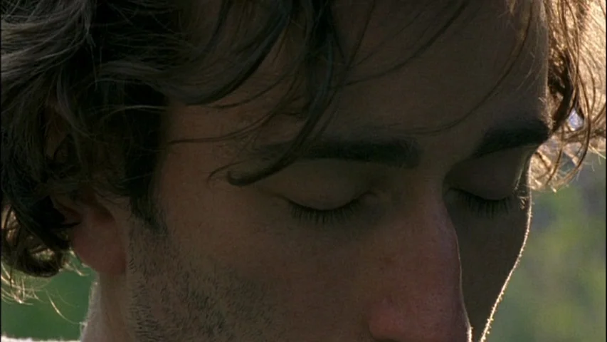 Old Joy (2006) movie still: closeup — An outdoor setting with natural light catching the side of a man's face. A man stands with his eyes closed…