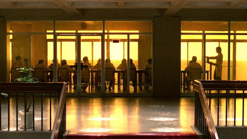 The Terrorizers (1986) movie still: wide — A modern building interior shows a row of figures seated behind a glass wall. The intense golden sunlight…