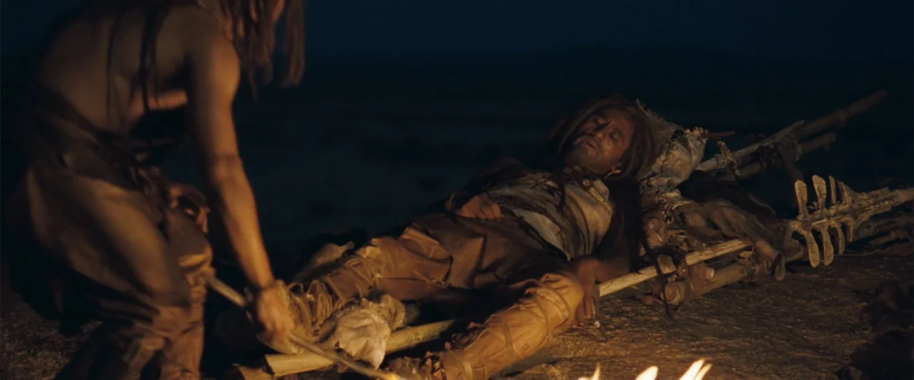 10,000 BC (2008) movie still: medium — A dark, expansive landscape at night with rocky ground and a flickering campfire. One man attends to another…