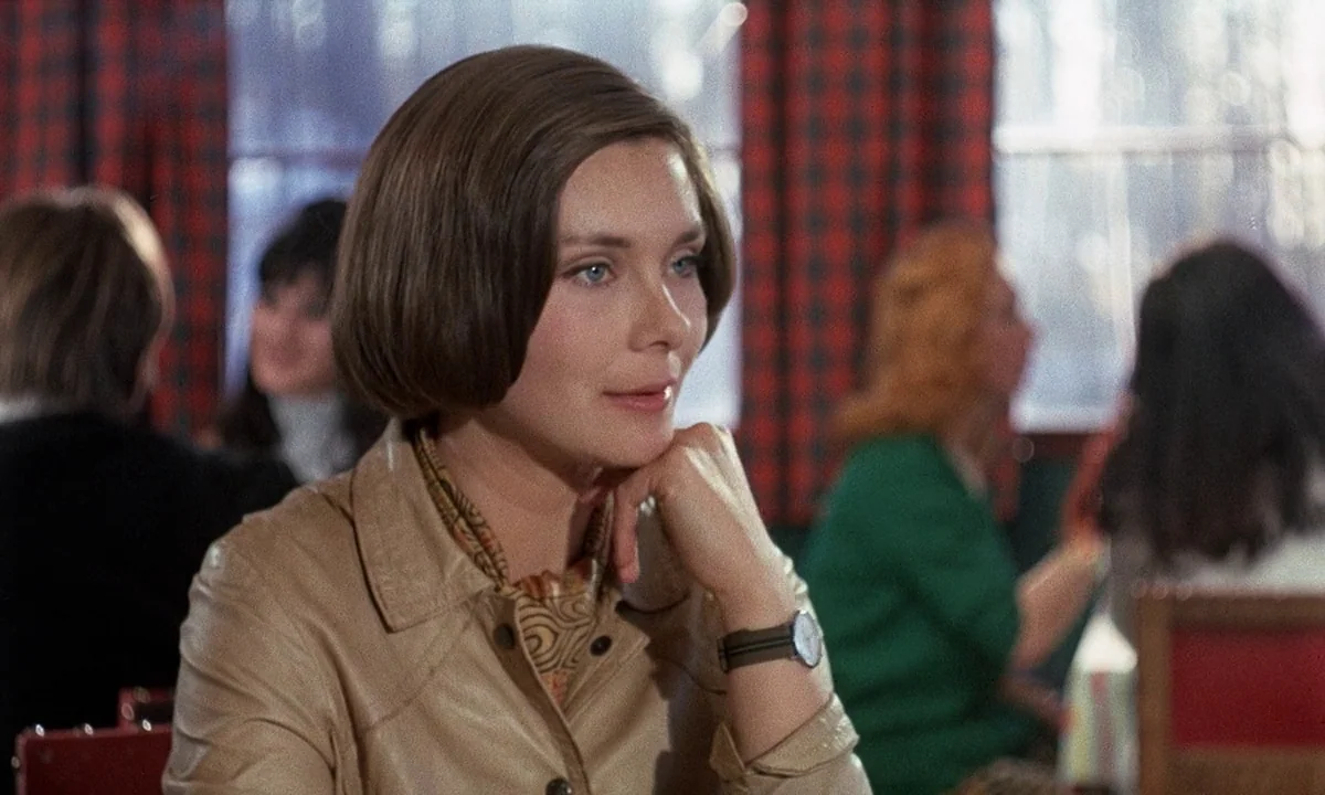 Belle De Jour (1967) movie still: medium — A woman sits in a restaurant booth with red checkered curtains in the background. She rests her chin on…
