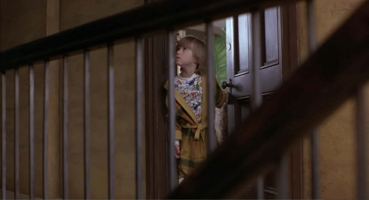 Bogus (1996) movie still: medium — A residential interior features a wooden staircase railing in the foreground. A young boy stands in an open…