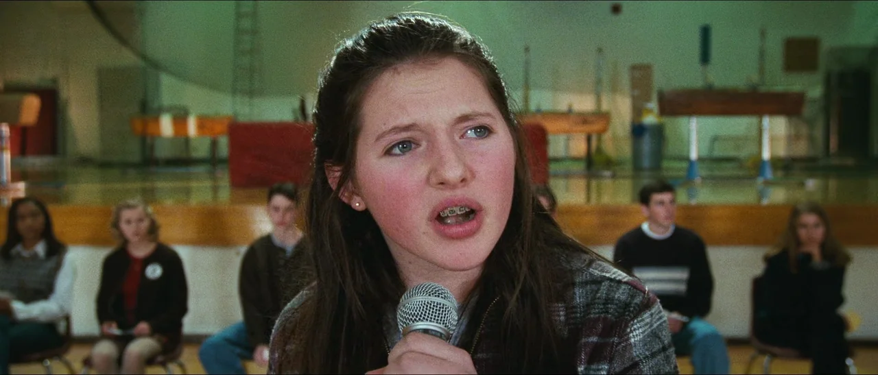Election (1999) movie still: closeup — A group of students sits in chairs in the background of a gymnasium. A teenage girl in the foreground holds a…