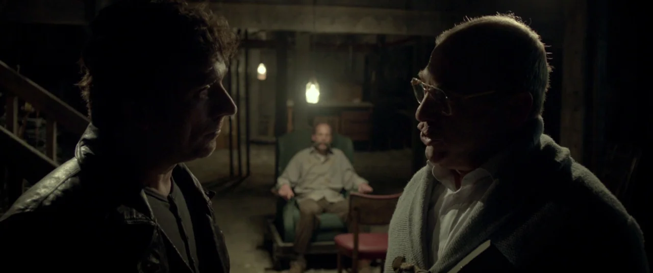 Big Bad Wolves (2013) movie still: medium — A dark, unfinished basement with exposed wooden joists and hanging light bulbs. Three men interact in…