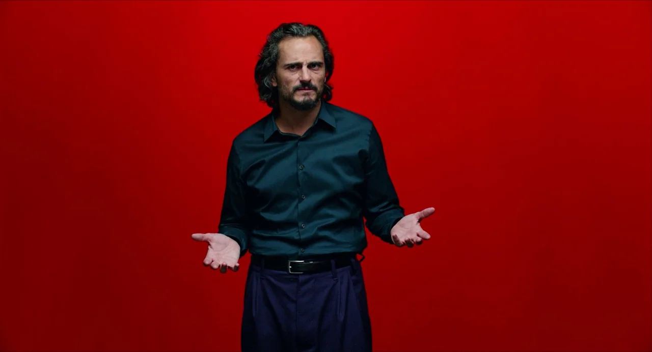 Pain and Glory (2019) movie still: medium — A man stands centered against a flat, solid red backdrop in a studio setting. The man holds his arms out…