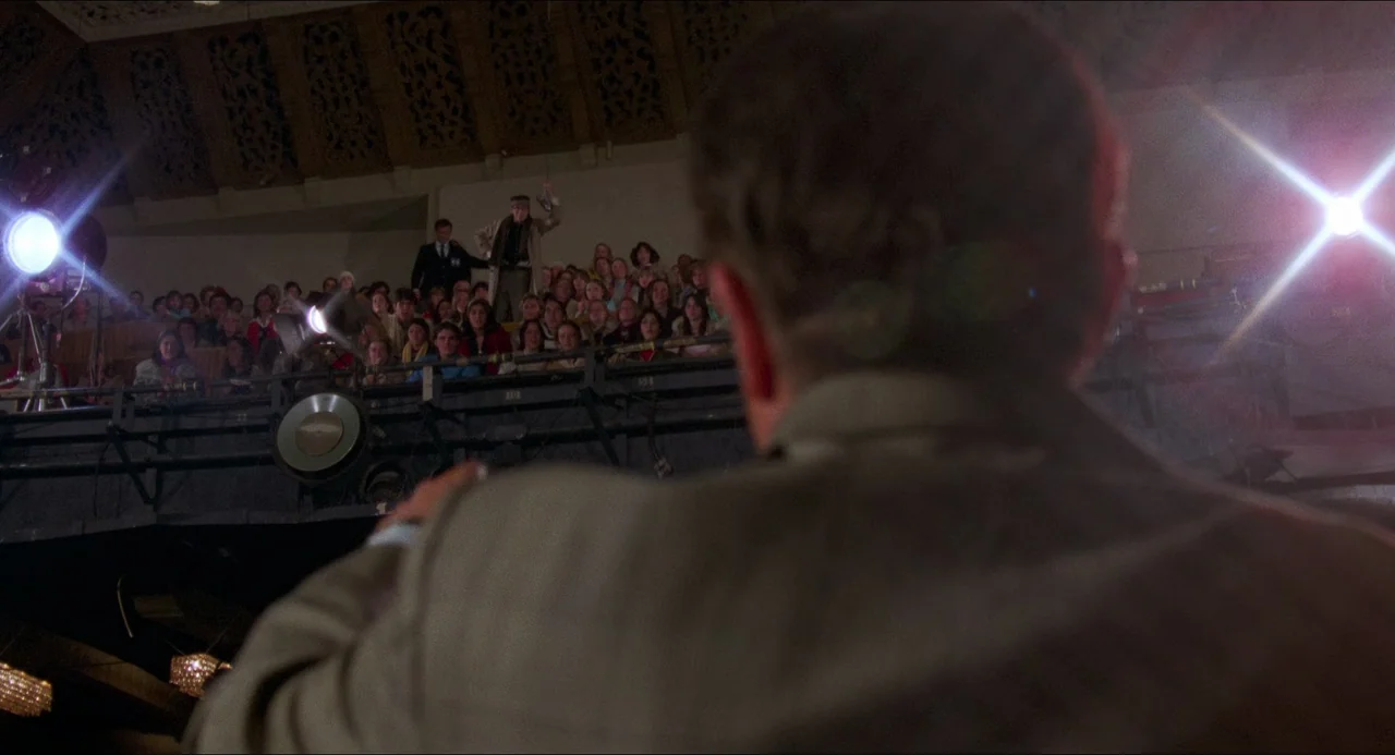 I Wanna Hold Your Hand (1978) movie still: medium — An auditorium with a large crowd gathered in the seating area viewed from behind. A person…