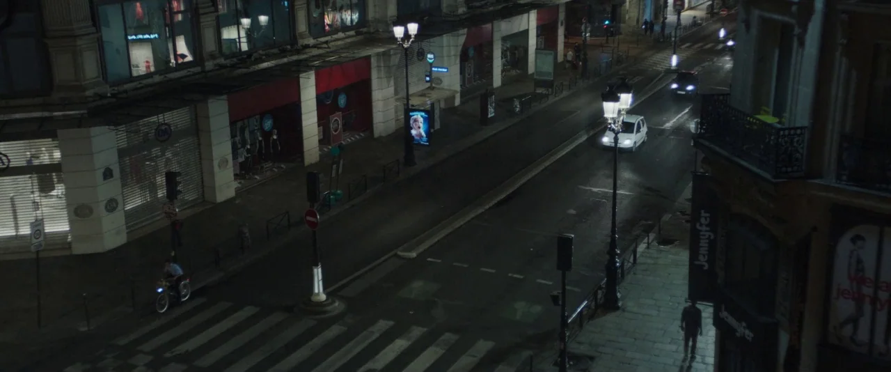 Nocturama (2016) movie still: establishing — A quiet city intersection at night features wet asphalt, multi-story buildings, and glowing…
