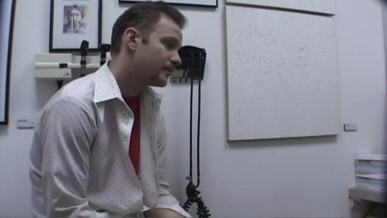 Super Size Me (2004) movie still: medium — A small clinical room with white walls and a framed artwork hanging behind a man. The man sits on a…