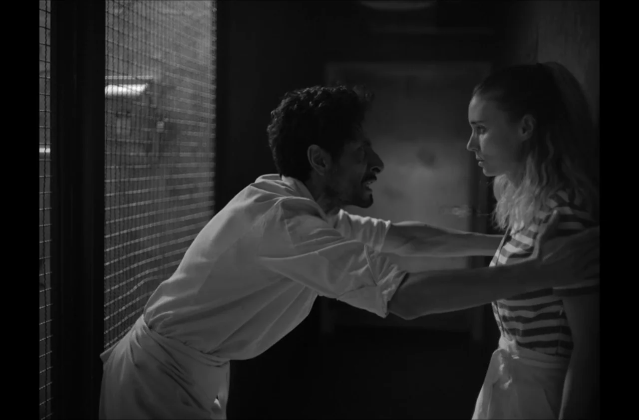 The Kitchen (2012) movie still: medium — A dimly lit narrow corridor features a chain-link fence panel along one wall. A man leans into a woman,…