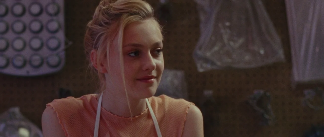 The Runaways (2010) movie still: closeup — A dimly lit room with pegboard walls and hanging plastic bags in the background. A young blonde woman…