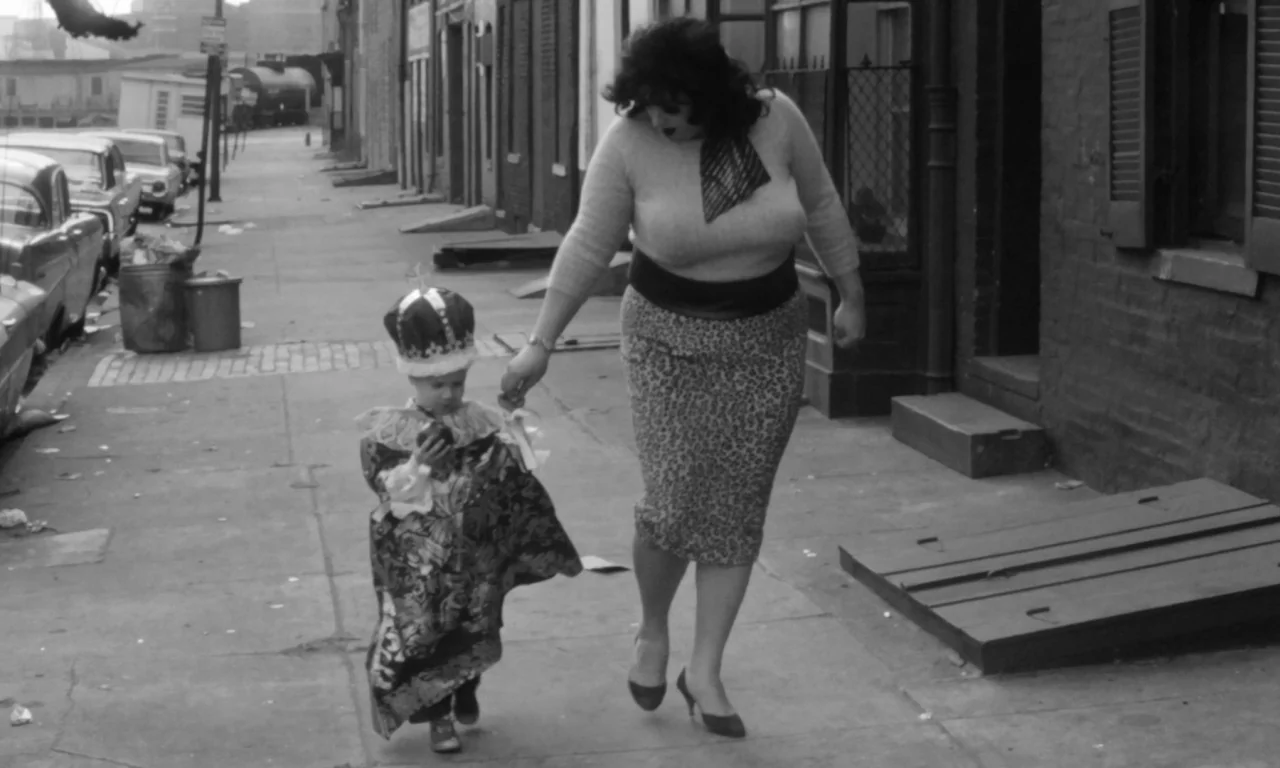 Multiple Maniacs (1970) movie still: fullbody — A sidewalk in an urban neighborhood lined with brick buildings and parked 1960s cars. A woman walks…