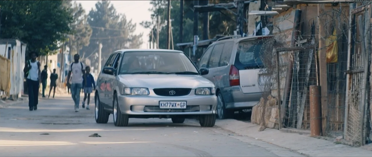 My Girlfriend's Father (2023) movie still: medium — A silver compact car drives slowly down a dusty road lined with makeshift structures.…