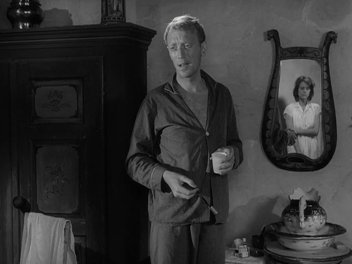 Through A Glass Darkly (1961) movie still: medium — A man in pajamas stands in a rustic room near a dark wooden cabinet. He holds a small cup while…