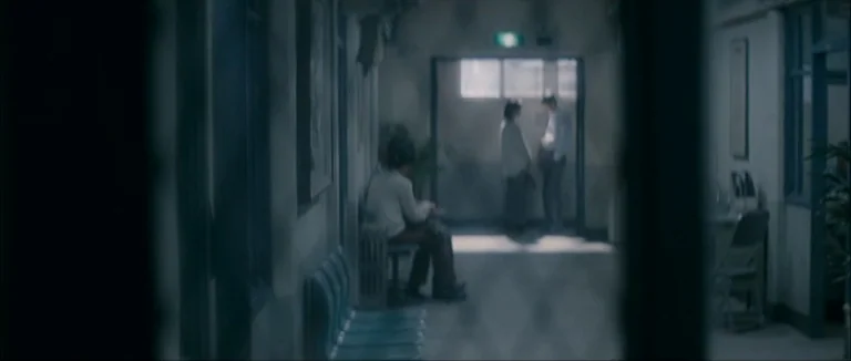 Mother (2009) movie still: wide — A long, dimly lit institutional hallway features rows of chairs and a doorway. A man sits alone while a couple…
