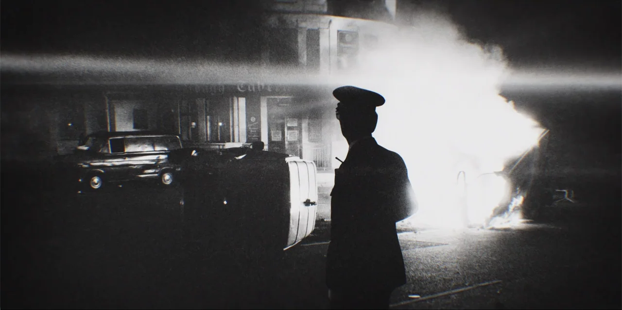 A Radical Life (2022) movie still: medium — An urban intersection features an overturned vehicle consumed by bright flames at night. A uniformed…