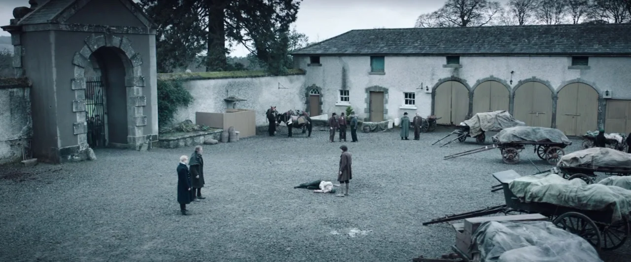 Black ’47 (2018) movie still: wide — A gravel-covered courtyard is enclosed by high stone walls and a stone gatehouse. Several men in dark coats…